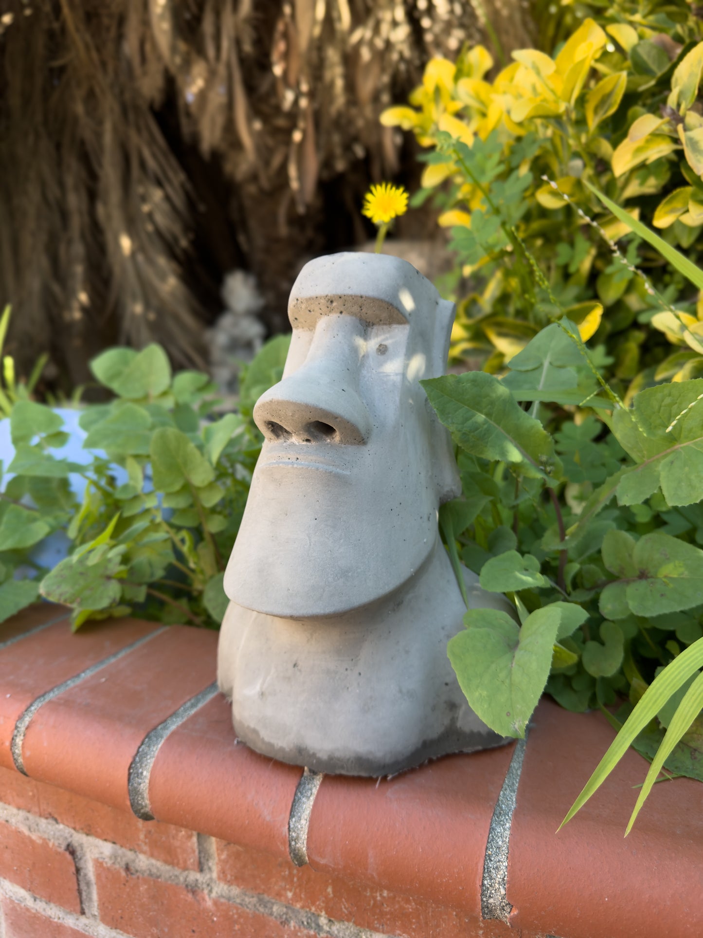 Concrete moai statue planter on a brick ledge next to an outdoor garden bed.