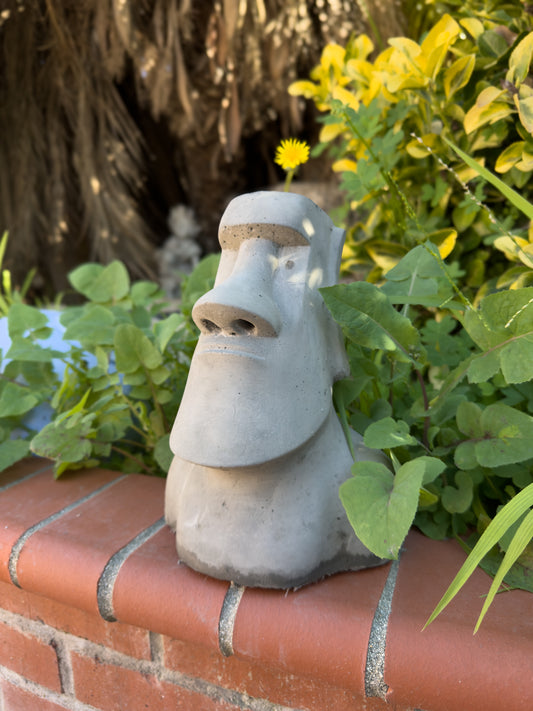 Concrete moai statue planter on a brick ledge next to an outdoor garden bed.