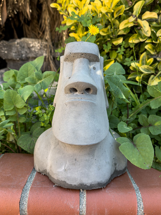 Concrete moai statue planter on a brick ledge next to an outdoor garden bed.